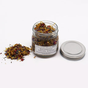 250g jar of dried lavender buds, rose petals, chamomile & calendula flowers spilling onto white marble with steaming bowl in soft spa setting