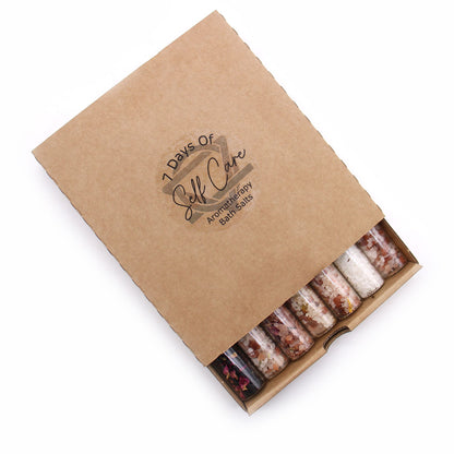 Elegant row of 7 coloured bath salt vials with dried flowers and essential oil droppers on marble tray, surrounded by candles and rose petals