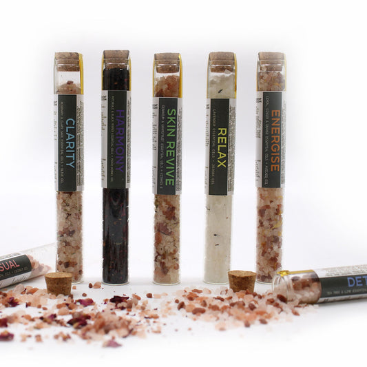 Elegant row of 7 coloured bath salt vials with dried flowers and essential oil droppers on marble tray, surrounded by candles and rose petals