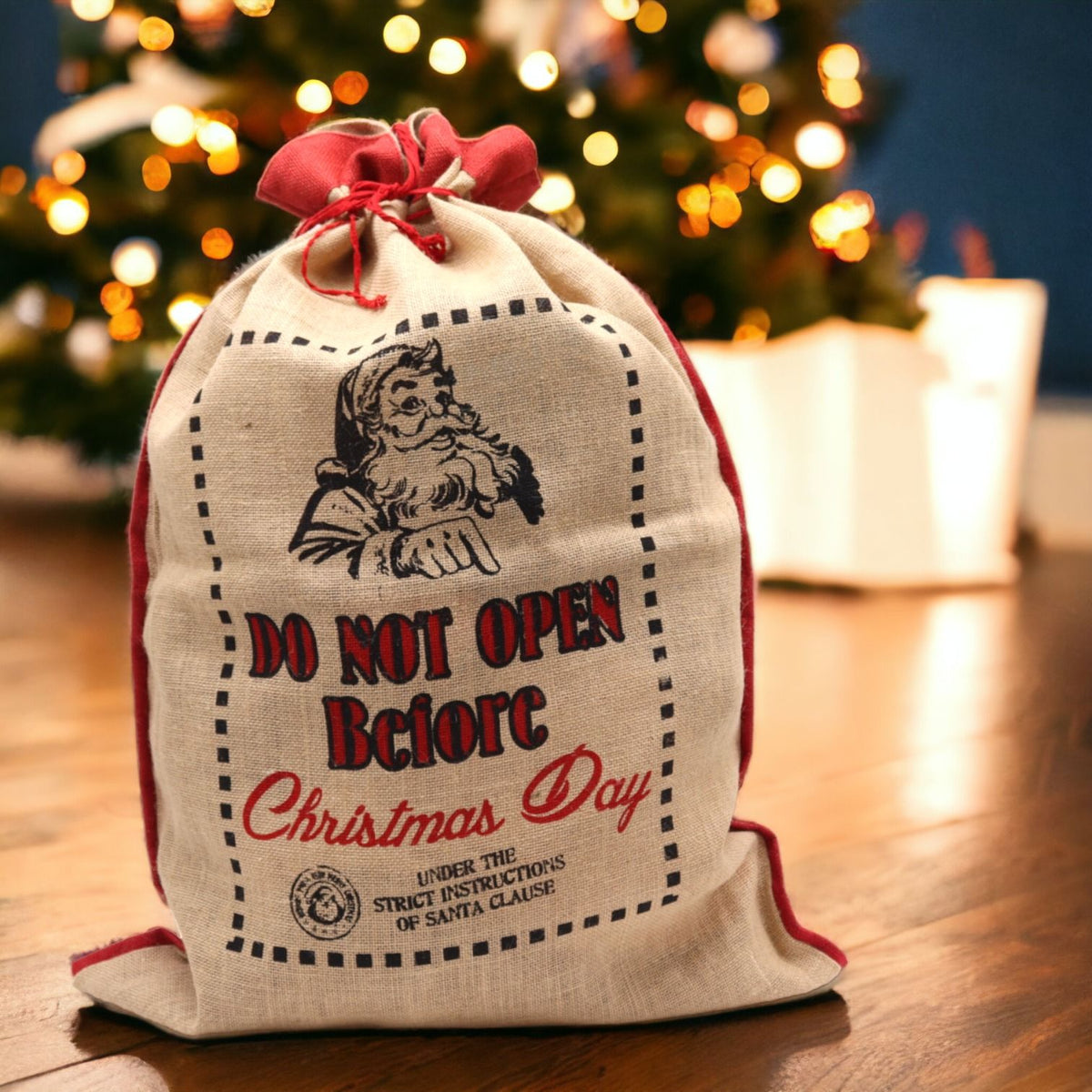 Large natural jute Christmas sack with bold “Do Not Open Before Christmas Day” print, overflowing with wrapped gifts, red ribbon tie and twinkling lights