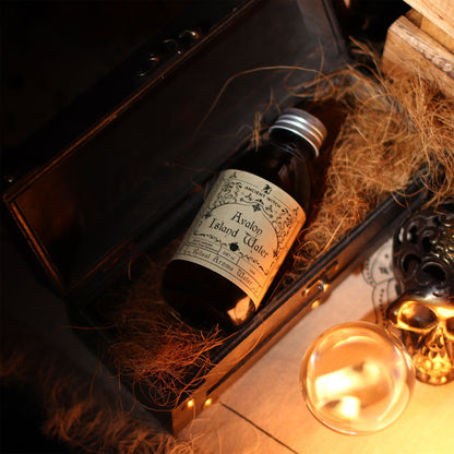 140ml amber bottle of Avalon Island ritual water with misty isle & nine priestesses label, surrounded by fresh lavender, roses and glowing candles