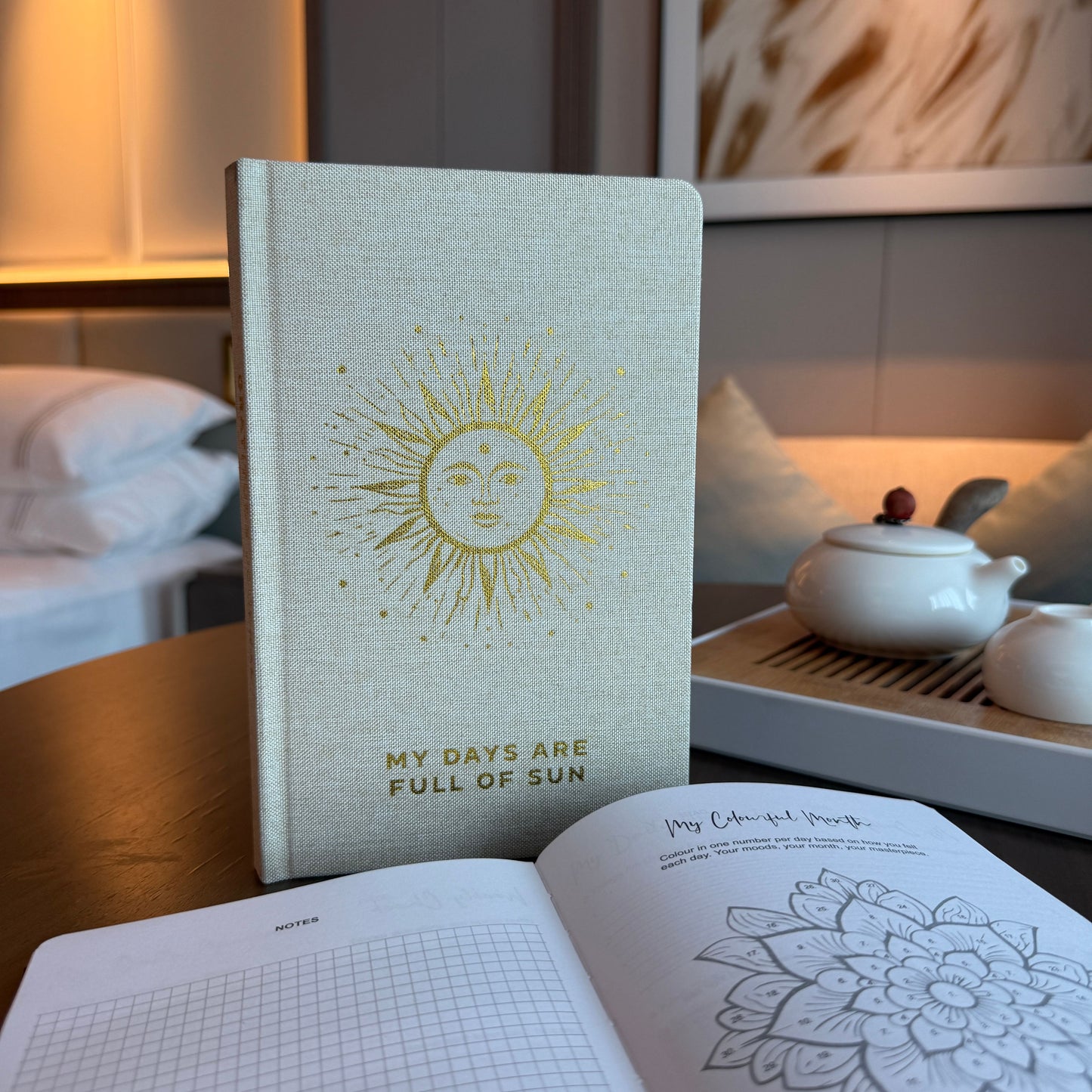 Notebook with sun design and text 'My days are full of sun' on a table with a teapot and open book.