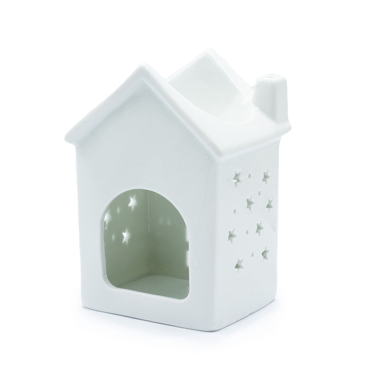 White ceramic cottage oil burner with glowing tealight inside, snow-dusted roof, heart door and warm window light on winter evening table with pine cones