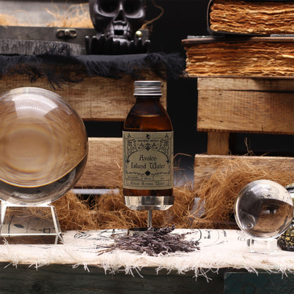 140ml amber bottle of Avalon Island ritual water with misty isle & nine priestesses label, surrounded by fresh lavender, roses and glowing candles