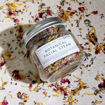 250g jar of dried lavender buds, rose petals, chamomile & calendula flowers spilling onto white marble with steaming bowl in soft spa setting