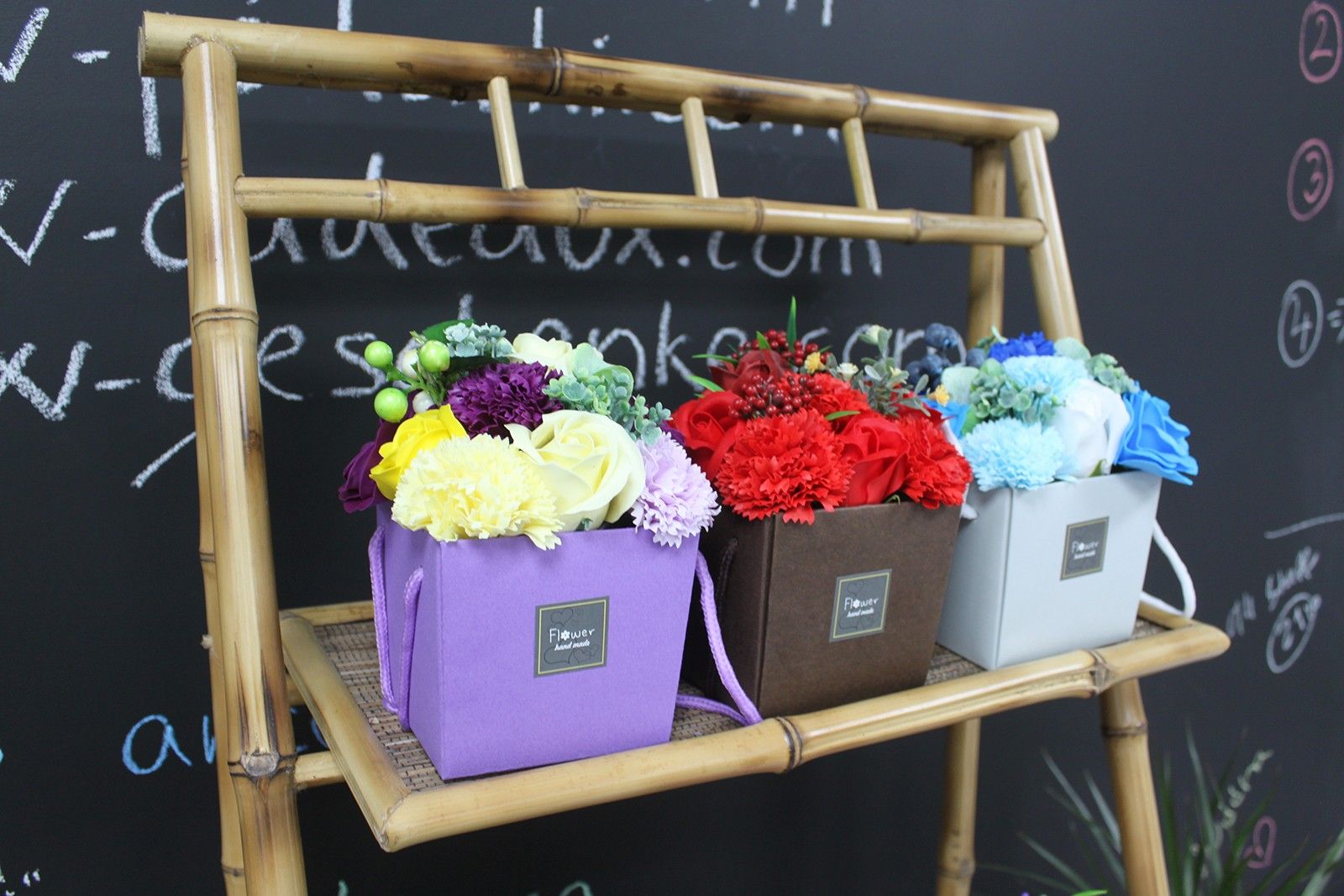 Three soap flower boxes on a bamboo stand against a chalkboard background