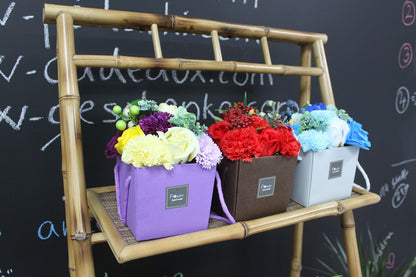 Three soap flower boxes on a bamboo stand against a chalkboard background