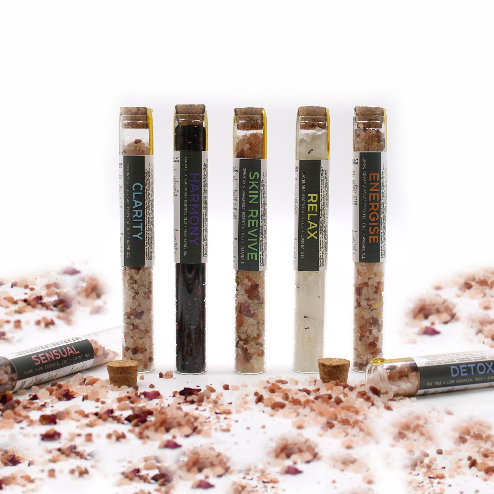 Elegant row of 7 coloured bath salt vials with dried flowers and essential oil droppers on marble tray, surrounded by candles and rose petals