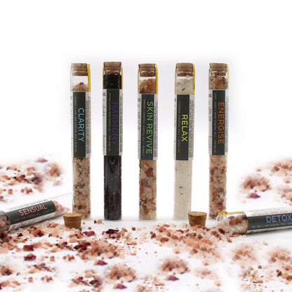 Elegant row of 7 coloured bath salt vials with dried flowers and essential oil droppers on marble tray, surrounded by candles and rose petals