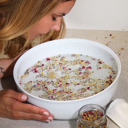 250g jar of dried lavender buds, rose petals, chamomile & calendula flowers spilling onto white marble with steaming bowl in soft spa setting