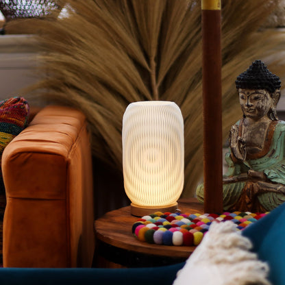 Decorative Modern Twisted New Wave USB lamp with textured honeycomb surface and sleek twisted silhouette, warm glow on 3rd brightness setting, placed on a wooden base on a table with a Buddha statue and colorful items in a cozy room setting.