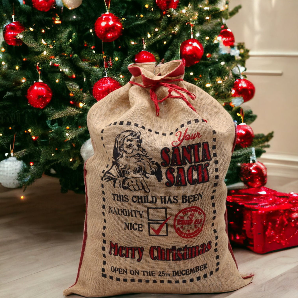 Large jute Christmas sack printed “This Child Has Been… Nice! ✓” with Santa and ticked Nice box, stuffed with toys and gifts under glowing tree