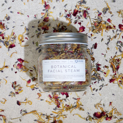 250g jar of dried lavender buds, rose petals, chamomile & calendula flowers spilling onto white marble with steaming bowl in soft spa setting