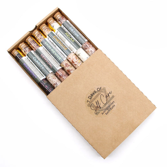 Elegant row of 7 coloured bath salt vials with dried flowers and essential oil droppers on marble tray, surrounded by candles and rose petals