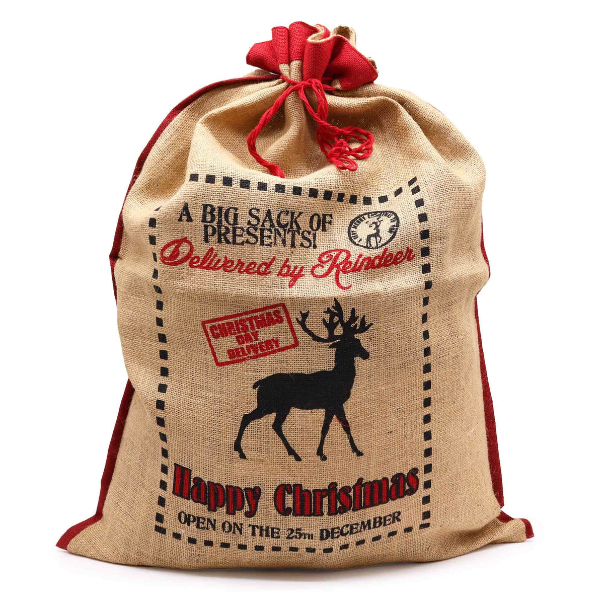 Large natural jute Christmas sack stamped “Delivered By Reindeer – Happy Christmas” filled with gifts, red ribbon tie, glowing tree lights in background