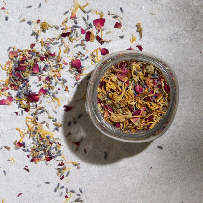 250g jar of dried lavender buds, rose petals, chamomile & calendula flowers spilling onto white marble with steaming bowl in soft spa setting