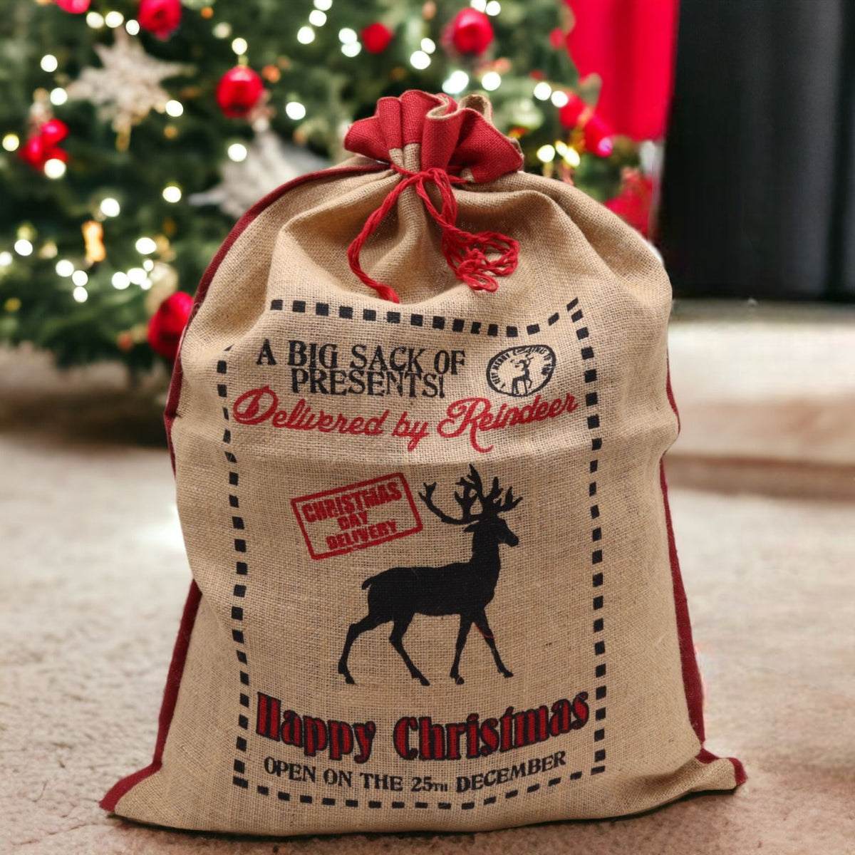 Large natural jute Christmas sack stamped “Delivered By Reindeer – Happy Christmas” filled with gifts, red ribbon tie, glowing tree lights in background
