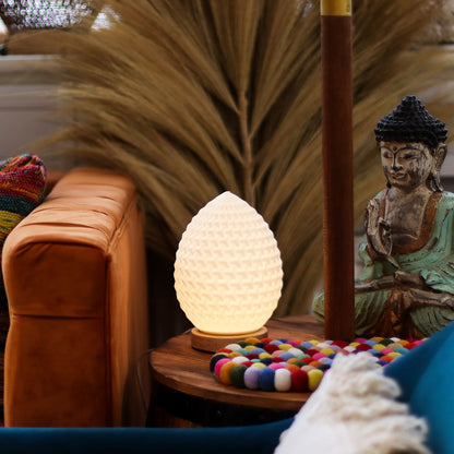 Decorative Teardrop New Wave USB lamp with textured honeycomb surface, warm glow on 3rd brightness setting, placed on a wooden base on a side table with a Buddha statue and colorful items in the background.