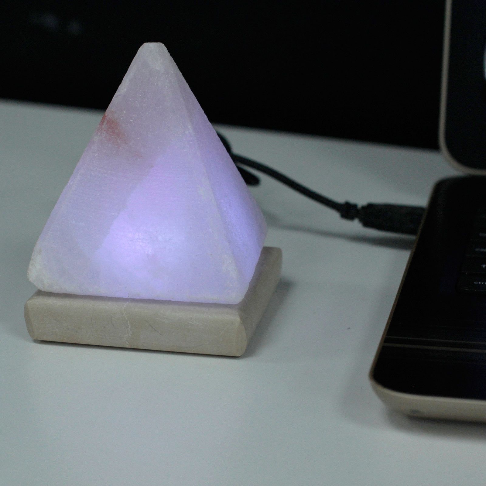 White pyramid Himalayan salt lamp cycling through rainbow colours, pink salt crystal glowing inside, USB cable, on minimalist white desk with laptop