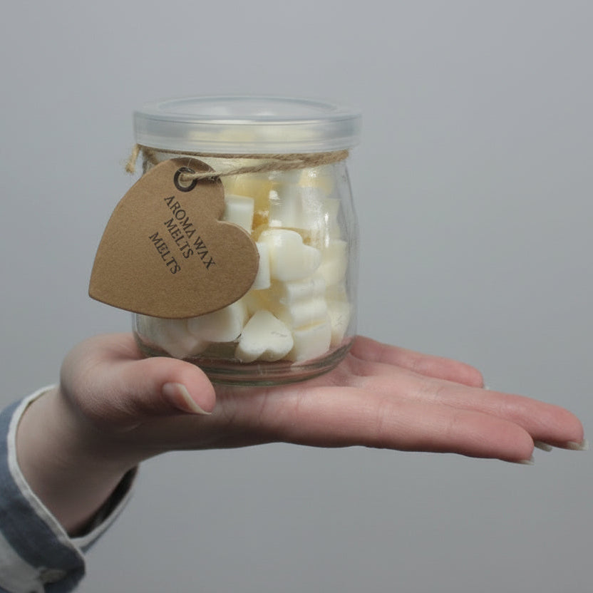 Hand holding a small glass jar filled with wax melts against a gray background