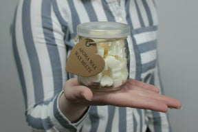 Hand holding a Glass jar of Nutmeg & Lemon Aroma Wax Melts with heart-shaped soy wax pieces, against a striped shirt background.